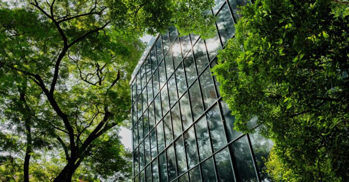 Modern glass building reflecting trees and sky in its windows, surrounded by lush greenery.