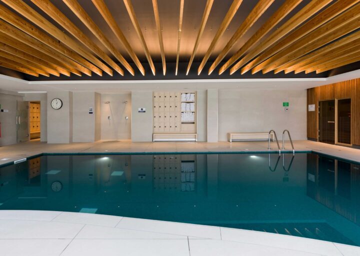 Indoor swimming pool area with a wooden ceiling, changing rooms, and lockers, featuring a clear blue pool and modern design elements.