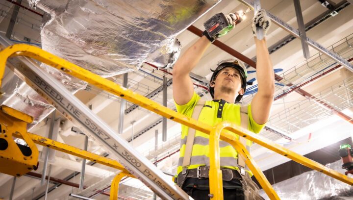 Construction worker in a high-visibility vest using a power tool while standing on a scaffold during a fit-out project.