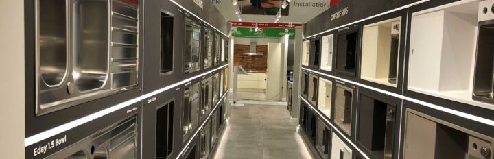 Display of various kitchen sink models arranged in a showroom aisle with stainless steel and composite options.